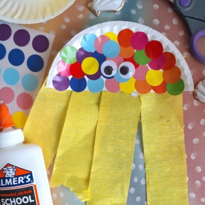 A colorful jellyfish craft made from a paper plate, rainbow dot stickers, googly eyes, and yellow crepe paper strips, surrounded by shells, scissors, glue, and @gluedtomycrafts watermark.