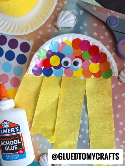 A colorful jellyfish craft made from a paper plate, rainbow dot stickers, googly eyes, and yellow crepe paper strips, surrounded by shells, scissors, glue, and @gluedtomycrafts watermark.