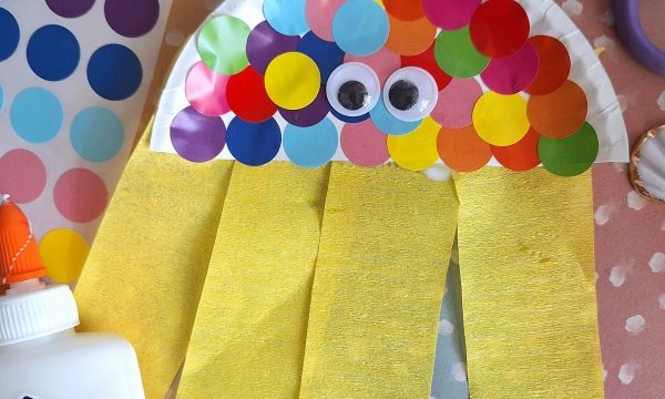 A colorful jellyfish craft made from a paper plate, rainbow dot stickers, googly eyes, and yellow crepe paper strips, surrounded by shells, scissors, glue, and @gluedtomycrafts watermark.