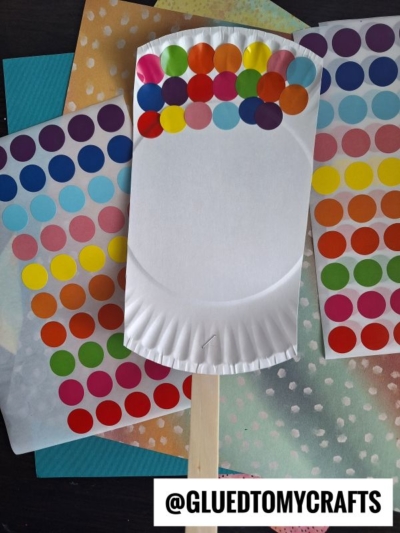 A white paper plate with colorful dot stickers arranged in rows is attached to a wooden stick, with sheets of similar colorful dot stickers in the background. @GLUEDTOMYCRAFTS is written at the bottom.