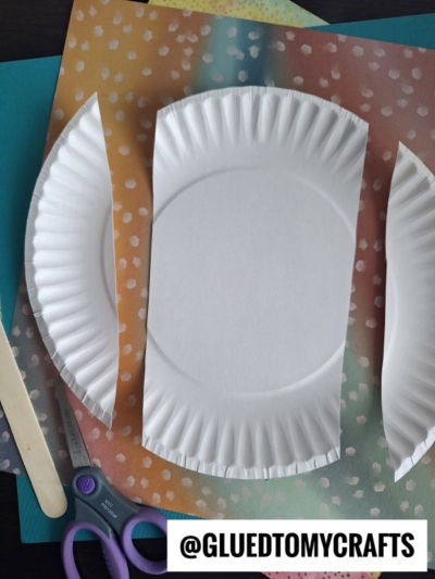 Two paper plates with sides cut off are placed on sheets of colorful, patterned paper. Scissors and a wooden craft stick are nearby. The image has @GLUETOMYCRAFTS text in the corner.