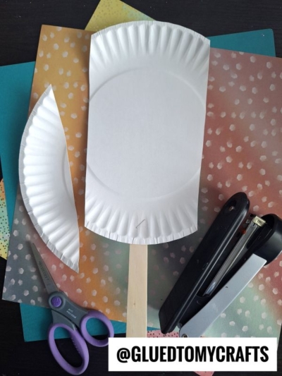 A paper plate cut and stapled onto a craft stick to resemble a popsicle, with scissors, a stapler, and colored papers in the background. The text @GLUEDTOMYCRAFTS is at the bottom.