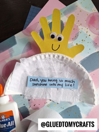 A handmade HANDPRINT FATHER'S DAY craft featuring a yellow handprint with googly eyes, attached to a white semicircle cloud. A note says, “Dad, you bring so much sunshine into my life!” with art supplies in the background.