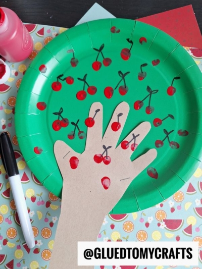 A green paper plate decorated with red cherry drawings and a cut-out paper hand glued on, with cherries drawn on the fingers. A black marker, pink paint, and patterned paper are nearby. @GLUEDTOMYCRAFTS is at the bottom.