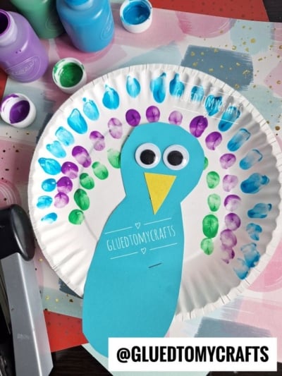 Create a vibrant paper plate fingerprint peacock with a blue paper body, yellow beak, googly eyes, and painted fingerprint feathers in blue, green, and purple. Paint bottles add extra color to this fun craft!.