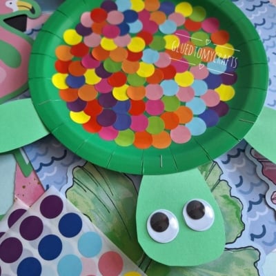 A green paper plate turtle craft with colorful round stickers on its back, green paper legs and head, and two large googly eyes. Craft materials and paper backgrounds are visible underneath.