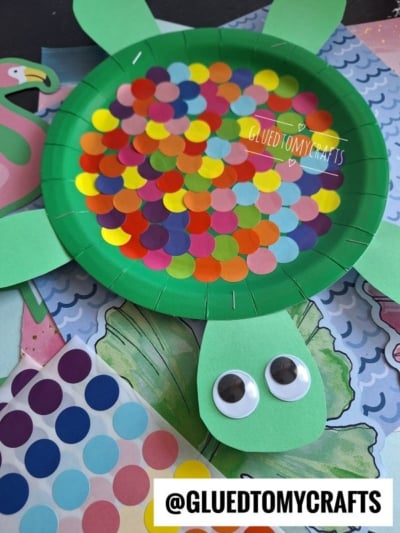 A green paper plate turtle craft with colorful round stickers on its back, green paper legs and head, and two large googly eyes. Craft materials and paper backgrounds are visible underneath.