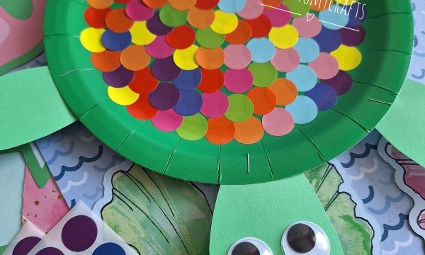 A green paper plate turtle craft with colorful round stickers on its back, green paper legs and head, and two large googly eyes. Craft materials and paper backgrounds are visible underneath.