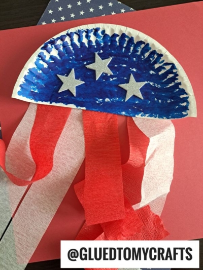 A paper plate painted blue with white stars and red and white streamers hanging down, this 4th of July craft resembles a patriotic jellyfish. The @GLUEDTOMYCRAFTS watermark is at the bottom.