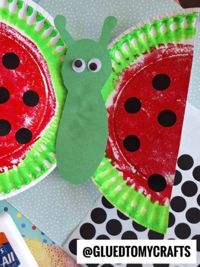 A green paper caterpillar with googly eyes is attached to two painted, wing-like paper plates decorated in red and green colors with black dots, resembling a paper plate watermelon butterfly. A @GLUEDTOMYCRAFTS label is at the bottom right.