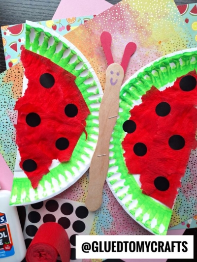 A butterfly craft made from two paper plate halves painted red with black dots, green edges, and attached to a popsicle stick body with pink antennae. Colorful paper and craft supplies are in the background.
