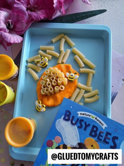 A tray with a bee-themed craft using pasta and orange Play-Doh, resembling a beehive. Perfect for story time sensory bin ideas, it features bee decorations, Play-Doh containers, flowers, the book Busy Bees, and the handle @GLUEDTOMYCRAFTS.