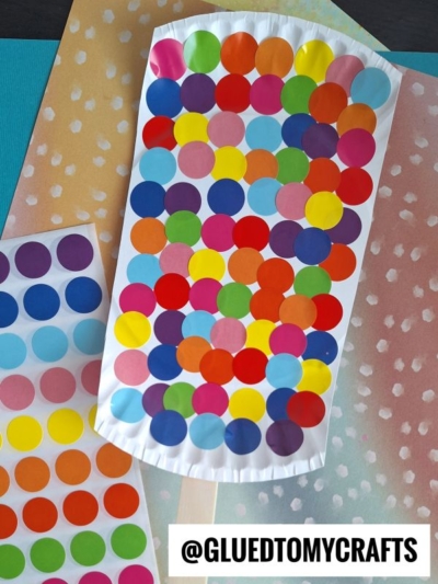 A colorful popsicle craft made from paper and covered with multicolored dot stickers, placed on pastel polka dot paper. A sheet of colorful dot stickers is beside it. @GLUEDTOMYCRAFTS is written at the bottom.