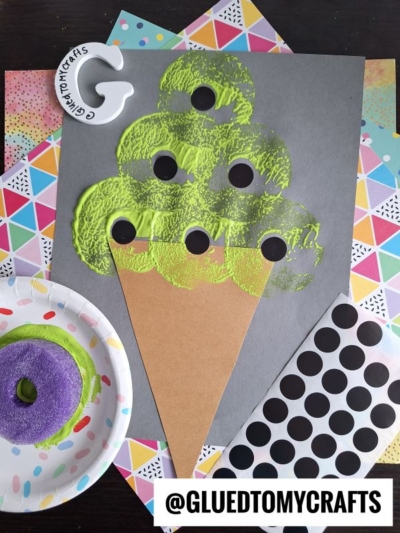A kids craft project featuring a green painted ice cream cone on gray paper, made with a paper cone, green paint, and black dot stickers—just like pool noodle stamped ice cream—surrounded by colorful patterned papers and crafting supplies.