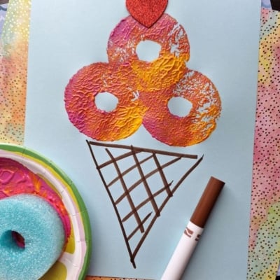 A piece of blue paper shows a colorful pool noodle stamped ice cream cone with orange, pink, and yellow scoops, topped with a red glitter heart. A brown marker and a paint-stained sponge rest nearby. The handle @GLUEDTOMYCRAFTS is at the bottom.