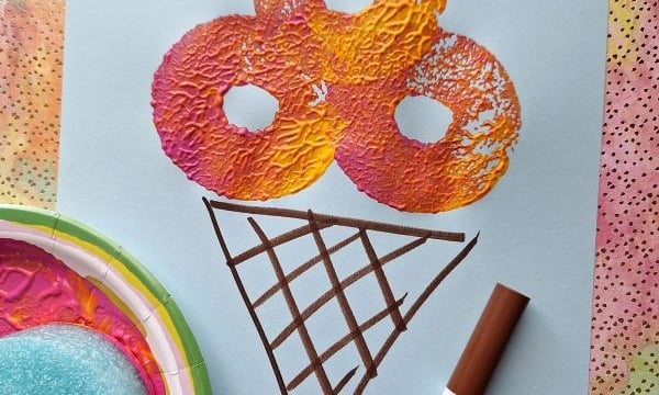 A piece of blue paper shows a colorful pool noodle stamped ice cream cone with orange, pink, and yellow scoops, topped with a red glitter heart. A brown marker and a paint-stained sponge rest nearby. The handle @GLUEDTOMYCRAFTS is at the bottom.