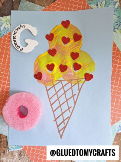 A colorful pool noodle stamped ice cream cone made from paint and red glitter hearts sits on blue paper. A pink sponge and a white letter “G” with writing are on the side. The handle @GLUEDTOMYCRAFTS is displayed at the bottom.
