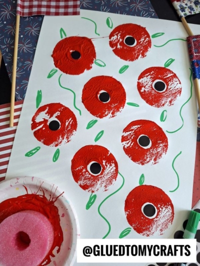 A sheet of paper displays red poppy flowers painted with black centers and green stems. A pink sponge dabbed in red paint is shown, along with craft supplies in the background. Text at the bottom reads @GLUEDTOMYCRAFTS.