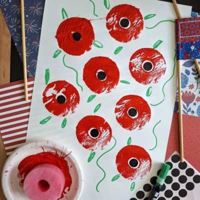 A white sheet features painted red poppies with black centers, green stems, and leaves. Nearby are patterned papers, two mini American flags, a pink sponge with red paint, a green marker, and the handle @GLUEDTOMYCRAFTS.