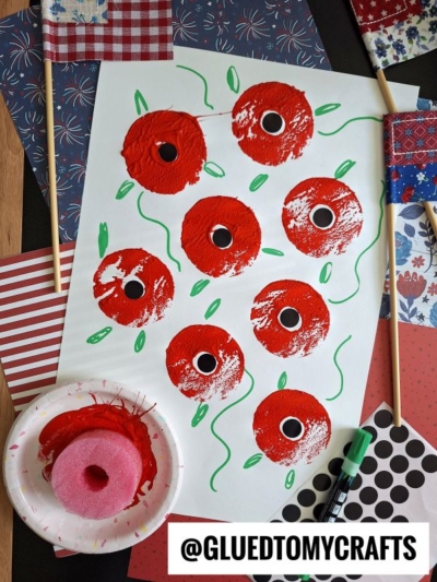 A white sheet features painted red poppies with black centers, green stems, and leaves. Nearby are patterned papers, two mini American flags, a pink sponge with red paint, a green marker, and the handle @GLUEDTOMYCRAFTS.