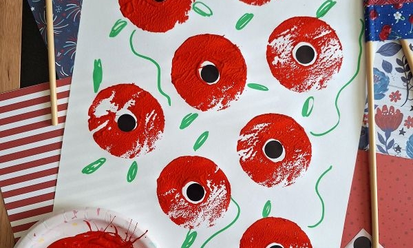 A white sheet features painted red poppies with black centers, green stems, and leaves. Nearby are patterned papers, two mini American flags, a pink sponge with red paint, a green marker, and the handle @GLUEDTOMYCRAFTS.