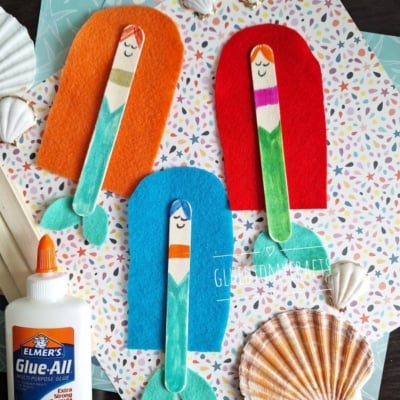 Three mermaid crafts made from colored popsicle sticks and felt are arranged on patterned paper with seashells and Elmer’s glue. @GluedToMyCrafts is displayed at the bottom right.
