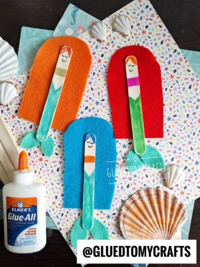 Three mermaid crafts made from colored popsicle sticks and felt are arranged on patterned paper with seashells and Elmer’s glue. @GluedToMyCrafts is displayed at the bottom right.