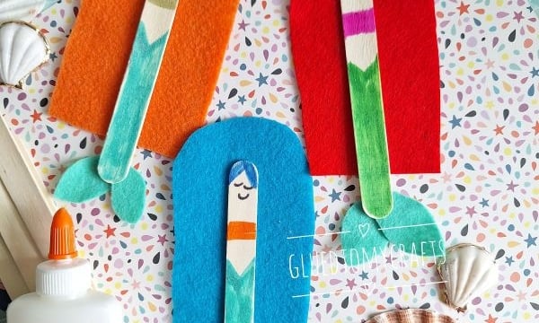 Three mermaid crafts made from colored popsicle sticks and felt are arranged on patterned paper with seashells and Elmer’s glue. @GluedToMyCrafts is displayed at the bottom right.