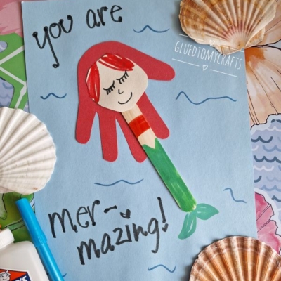 A handmade mermaid craft made from colored paper and a popsicle stick is on a blue sheet with You are mer-mazing! written on it. The scene is decorated with seashells, markers, glue, and @GLUEDTOMYCRAFTS.