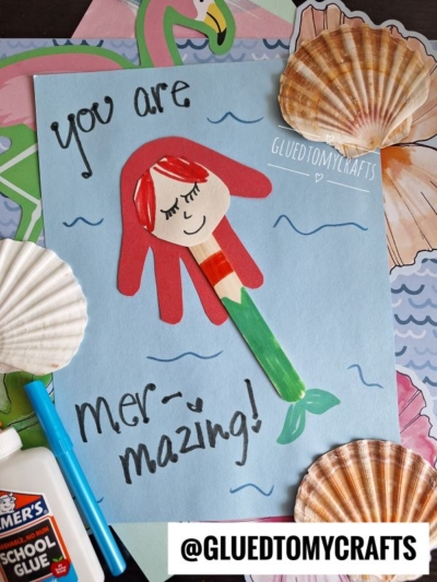 A handmade mermaid craft made from colored paper and a popsicle stick is on a blue sheet with You are mer-mazing! written on it. The scene is decorated with seashells, markers, glue, and @GLUEDTOMYCRAFTS.