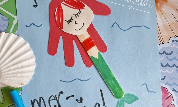 A handmade mermaid craft made from colored paper and a popsicle stick is on a blue sheet with You are mer-mazing! written on it. The scene is decorated with seashells, markers, glue, and @GLUEDTOMYCRAFTS.