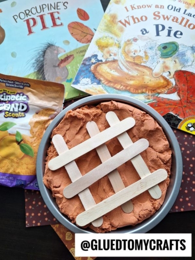 A round pan with brown kinetic sand and craft sticks arranged like a pie crust, placed near pie-themed children's books and a packet of Kinetic Sand—perfect for fun kinetic sand play ideas. The handle @GLUEDTOMYCRAFTS is on the bottom of the image.