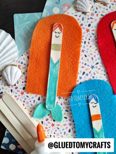 A craft project featuring a mermaid made from a popsicle stick, orange felt for hair, blue felt for the tail, and drawn facial features, surrounded by seashells and patterned paper.