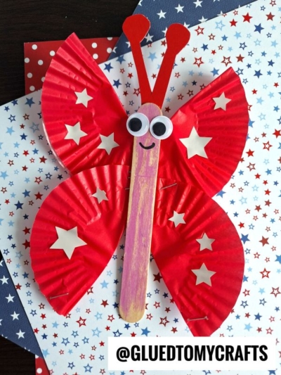 A butterfly craft made from red cupcake liners, a wooden stick decorated with a smile and googly eyes, and white star stickers, displayed on star-patterned paper—perfect for those seeking fun insect craft ideas. The handle @GLUEDTOMYCRAFTS is at the bottom.