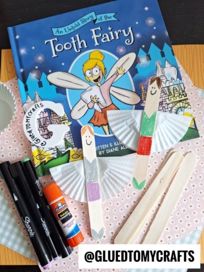 A children’s book titled “An Unfold Story of the Tooth Fairy” is surrounded by craft supplies: markers, glue stick, wooden craft sticks decorated as fairies, cupcake liners, and a @GLUEDTOMYCRAFTS handle.