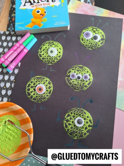 A black paper displays green, textured monster faces made with paint and googly eyes. Colorful markers, a book, and painting tools surround the craft materials. The Instagram handle @GLUETOMYCRAFTS is visible.