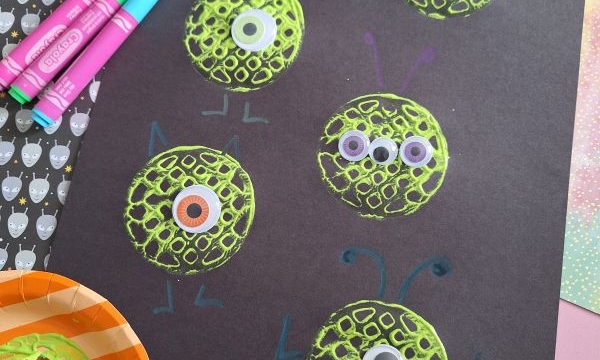 A black paper displays green, textured monster faces made with paint and googly eyes. Colorful markers, a book, and painting tools surround the craft materials. The Instagram handle @GLUETOMYCRAFTS is visible.