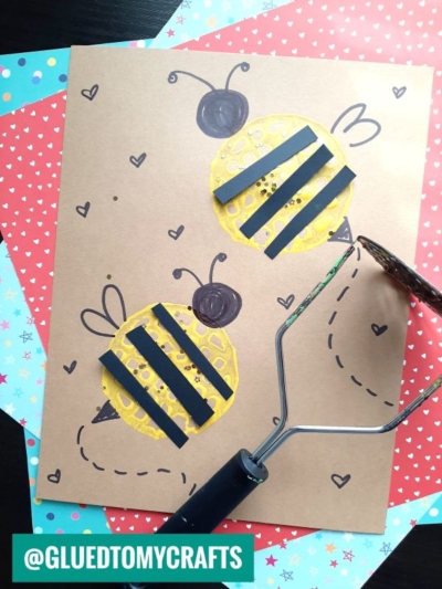 Craft paper with two potato masher stamped bees made from yellow paint circles and black paper strips. Drawn wings and dots decorate the bees. A textured roller and colorful papers are nearby. Text reads @GLUEDTOMYCRAFTS.