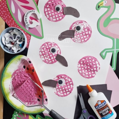 A craft setup featuring five pink bird prints on paper, made using a painted bubble wrap stamp. Nearby are googly eyes, scissors, glue, paper cutouts, and a flamingo illustration. The handle @GLUEDTOMYCRAFTS is shown.