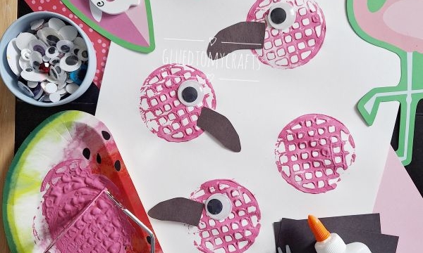 A craft setup featuring five pink bird prints on paper, made using a painted bubble wrap stamp. Nearby are googly eyes, scissors, glue, paper cutouts, and a flamingo illustration. The handle @GLUEDTOMYCRAFTS is shown.