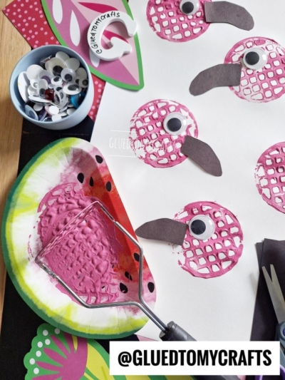 A kids craft scene with pink paint on a paper plate, a potato masher used for stamping, googly eyes, paper cutouts, and printed shapes resembling flamingos with black beaks on white paper.