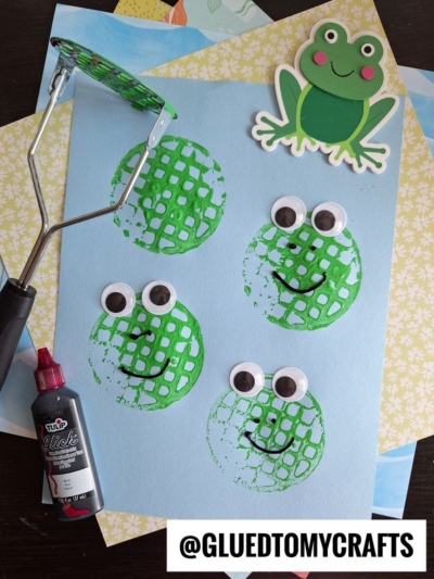 A blue paper showcases a potato masher stamped craft featuring four green frog faces with googly eyes and drawn-on smiles. Nearby are a bottle of black paint, a potato masher, and a cheerful cartoon frog.