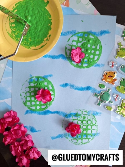 A creative children's potato masher stamped craft with green painted lily pads, pink paper flowers, and blue waves on paper. A potato masher, floral stickers, and pink flowers are beside the artwork. @GLUEDTOMYCRAFTS is at the bottom.