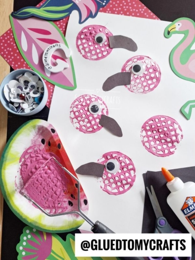 Art supplies and paper crafts on a table show pink flamingos made from stamped circles, googly eyes, and paper beaks, surrounded by scissors, glue, a potato masher with pink paint, and tropical-themed cutouts.