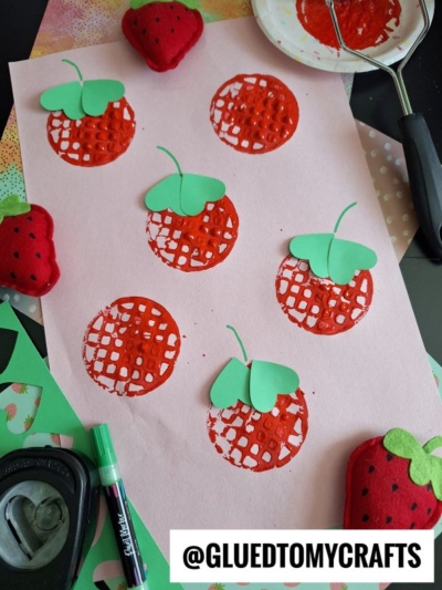 A pink sheet of paper with red strawberry prints, each topped with a green paper leaf. Craft tools, paper scraps, and plush strawberries surround the sheet. A label reads “@GLUEDTOMYCRAFTS.”.