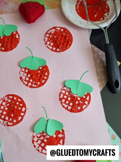 Pink paper with red circular prints resembling strawberries, made using a potato masher and paint. Green paper leaves are glued on top of each print. A red felt strawberry and crafting supplies are nearby. @GLUEDTOMYCRAFTS watermark.