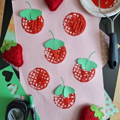 A pink sheet of paper with stamped red strawberries, some topped with green paper leaves, surrounded by crafting tools, green paper cutouts, a red paint roller, and two plush strawberry toys. @GLUEDTOMYCRAFTS is labeled at the bottom.