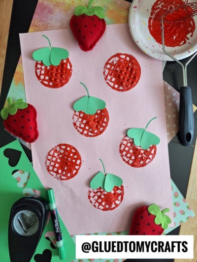 A pink sheet of paper with stamped red strawberries, some topped with green paper leaves, surrounded by crafting tools, green paper cutouts, a red paint roller, and two plush strawberry toys. @GLUEDTOMYCRAFTS is labeled at the bottom.
