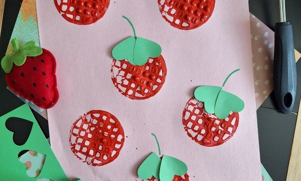 A pink sheet of paper with stamped red strawberries, some topped with green paper leaves, surrounded by crafting tools, green paper cutouts, a red paint roller, and two plush strawberry toys. @GLUEDTOMYCRAFTS is labeled at the bottom.