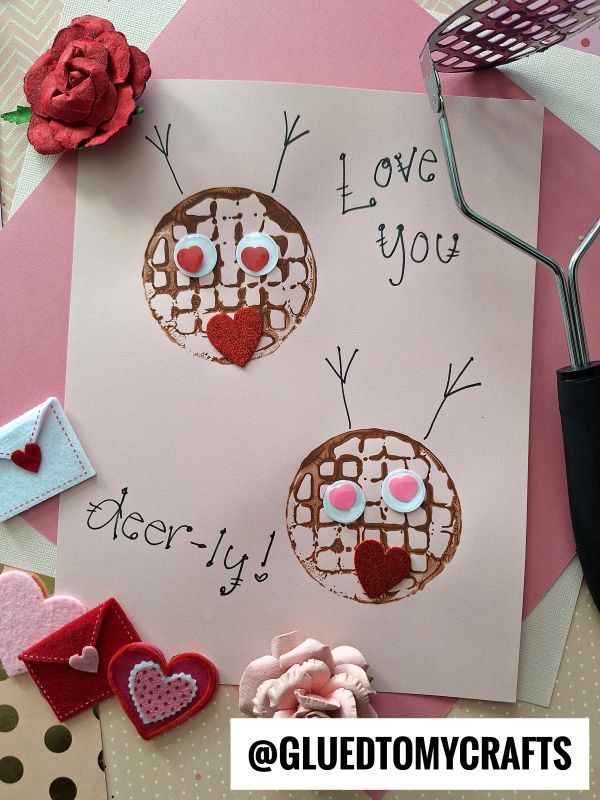 A handmade Valentine’s card features two potato masher stamped reindeer with googly eyes and red felt hearts for noses, surrounded by decorative hearts and a Love you deer-ly! message.